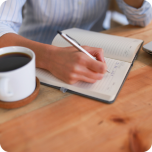 image of someone writing a list in a notebook, seated at a table with a coffee on the table